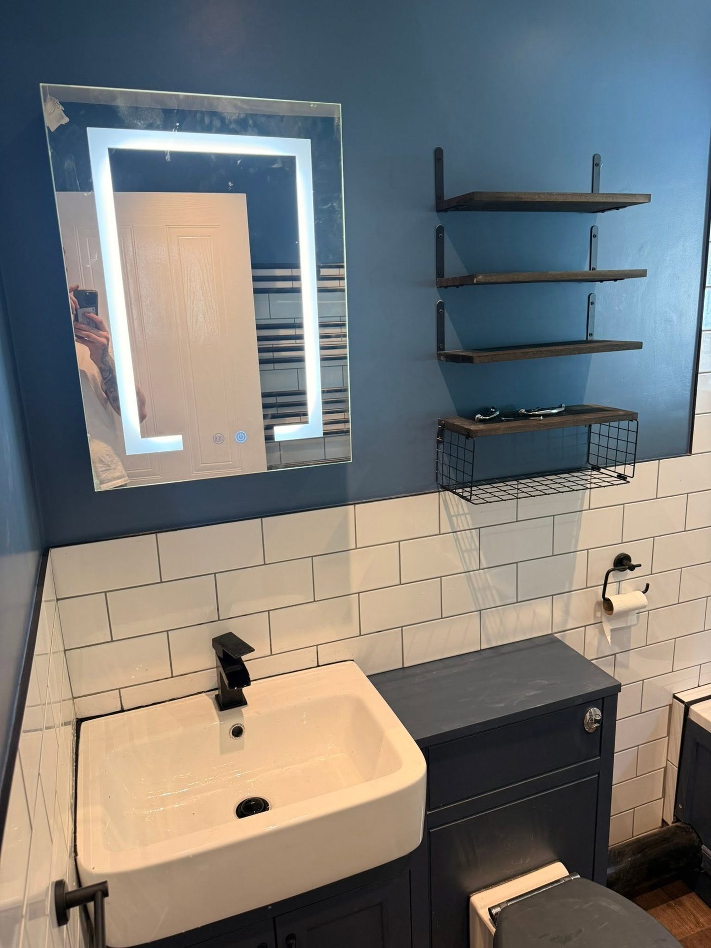 Grey vanity unit with a matt black square mixer tap, backlit LED mirror, white metro tiling and blue walls in a bathroom refitted by JY Plumbing & Heating